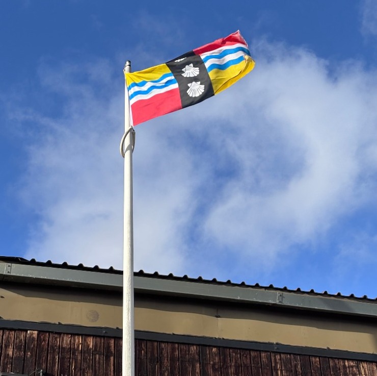 The Bedfordshire flag raised above Biggleswade rugby club. The flag's yellow and red quadrants are taken from the arms of the Beauchamp family, powerful in the county after the Norman conquest of England and constructors of Bedford Castle. The blue and white wavy lines symbolise the Great Ouse, which flows through the county. The three shells are taken from the coat of arms of the Duke of Bedford.