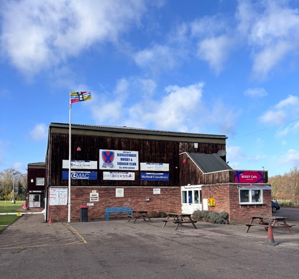 The Bedfordshire flag raised above Biggleswade rugby club. The flag's yellow and red quadrants are taken from the arms of the Beauchamp family, powerful in the county after the Norman conquest of England and constructors of Bedford Castle. The blue and white wavy lines symbolise the Great Ouse, which flows through the county. The three shells are taken from the coat of arms of the Duke of Bedford.